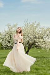 Fototapeta na wymiar A young beautiful blonde in a long white dress poses near a cherry blossom in the garden, a spring picturesque landscape