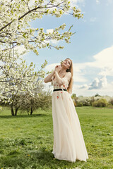 A young beautiful blonde in a long white dress poses near a cherry blossom in the garden, a spring picturesque landscape