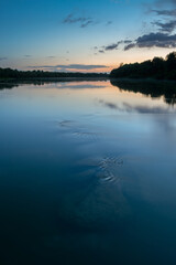 Underwater boulders in river at dusk, landscape of Sava river with forest silhouette on shore and clouds reflecting in water