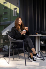 Dreamy girl with closed eyes holding coffee drink, legs crossed. Attractive young woman relaxing outdoors in a cafe.