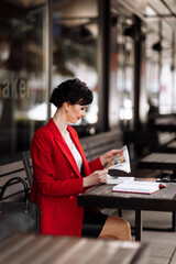 A attractive businesswoman in corporate outfit red blazer holding morning newspaper while sitting on cafe terrace. Manager is reading the latest world news during a coffee break at work