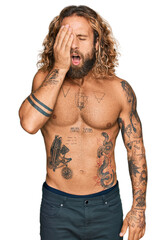 Handsome man with beard and long hair standing shirtless showing tattoos yawning tired covering half face, eye and mouth with hand. face hurts in pain.