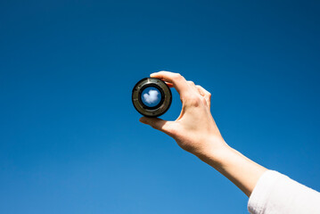Woman hand holding a photographic lens aiming at a white cloud in blue sky. Freedom, success, pursue and reach goals concept. - obrazy, fototapety, plakaty