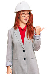 Young redhead woman wearing architect hardhat smiling with happy face looking and pointing to the side with thumb up.