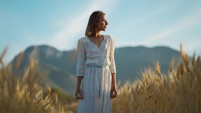 Young charming woman standing in golden field and looking away to sunset lights. Girl in white long dress in wheat field with mountains behind