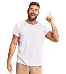 Young hispanic man wearing casual white tshirt pointing finger up with successful idea. exited and happy. number one.