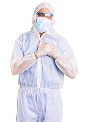 Young hispanic man wearing doctor protection coronavirus uniform and medical mask smiling with hands on chest with closed eyes and grateful gesture on face. health concept.