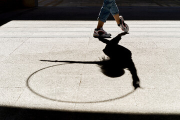 Crop legs and shadow of anonymous slim female in jeans and sneakers performing dance movements with hula hoop on paved street in sunny day