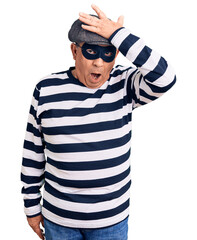 Senior handsome man wearing burglar mask and t-shirt surprised with hand on head for mistake,...