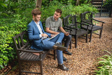 Two businessman or gay friend partner sitting at chair in park while using digital tablet and paper...