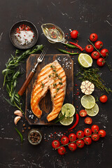 Grilled salmon steak on vintage wooden board with fork, olive oil, vegetables, herbs and spices on dark concrete background. Top view, copy space, flat lay