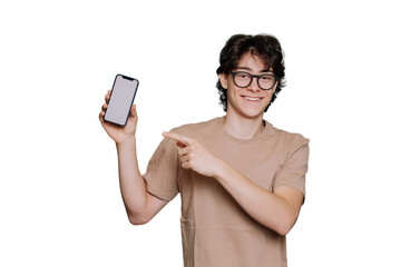 Happy caucasian boy in glasses, beige t-shirt holds phone toothy smiles points at blank screen of smartphone by index finger stands against transparent background. Smart Italian teen loves gadgets.