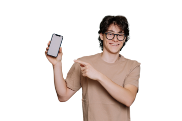 Happy caucasian boy in glasses, beige t-shirt holds phone toothy smiles points at blank screen of smartphone by index finger stands against transparent background. Smart Italian teen loves gadgets.