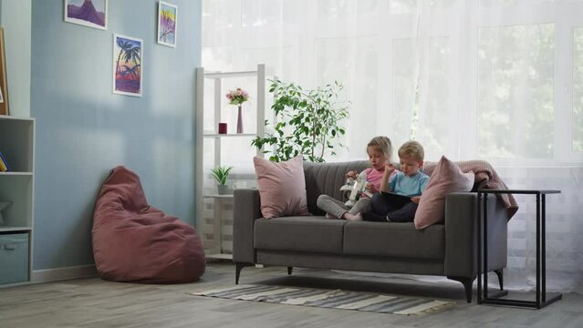 Preschooler girl holds airplane toy while toddler boy looks in tablet. Siblings sit on sofa near big window watching educational lessons on gadget