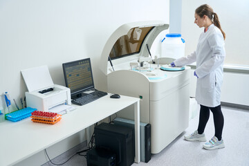 Young employee in overalls stands in front modern biochemical analyzer
