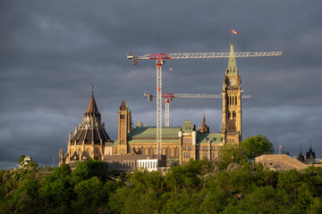View of Canadian Parliament Building at a Parliament hill by Ottawa river lit up at sunset hour, heavy storm clouds in the background.