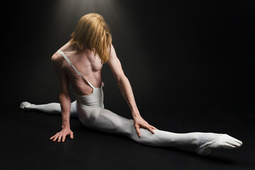 Fototapeta na wymiar Young athletic professional ballet dancer with a bare torso and white dance tights is in perfect shape, performing and doing splits over a black background.