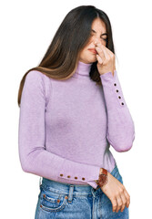 Young beautiful teen girl wearing turtleneck sweater smelling something stinky and disgusting, intolerable smell, holding breath with fingers on nose. bad smell - obrazy, fototapety, plakaty