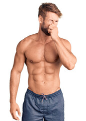 Young caucasian man standing shirtless smelling something stinky and disgusting, intolerable smell,...
