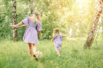 Naklejka na ściany i meble mother and daughter in short lilac dresses run along a summer meadow with green grass and trees. A small red dog runs in front of them.