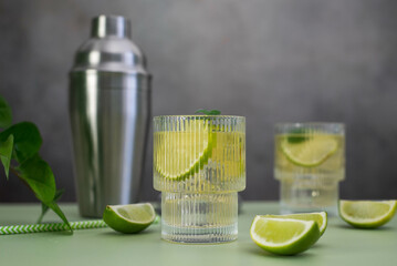 two fresh cold glasses of mahito with lime and ice and shaker on a gray-green background.