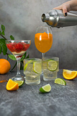 hand woman is holding shaker and pouring coctail in glass. assorted cocktails: two glasses of mojito, orange cocktail, cocktail with strawberries, fruits and shaker on a gray background.