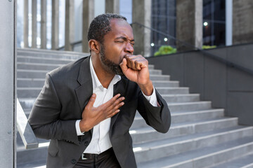 Cough attack on the street. Senior African American businessman standing and coughing suffocatingly, choking, covering his mouth with his hand, holding his chest, having difficulty breathing. - Powered by Adobe