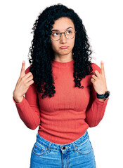 Young hispanic woman with curly hair wearing glasses pointing up looking sad and upset, indicating direction with fingers, unhappy and depressed.