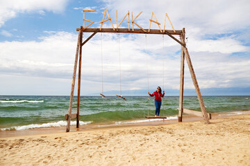 A woman swings on a wooden swing on the sandy beach of Lake Baikal. Summer vacation. Barguzinsky Bay, Republic of Buryatia.