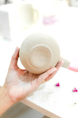 hand of a young craftswoman holding a clay mug she has made in her workshop. her work table is cluttered with many tools.