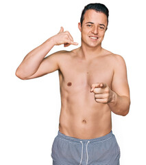 Fototapeta premium Handsome young man wearing swimwear shirtless smiling doing talking on the telephone gesture and pointing to you. call me.