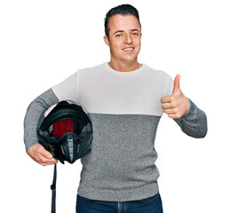 Handsome young man holding motorcycle helmet smiling happy and positive, thumb up doing excellent...
