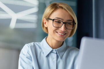 Beautiful businesswoman close up at workplace working with laptop, female worker at work inside modern office, blonde looking at laptop screen and smiling satisfied with achievement results.