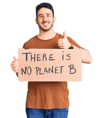 Young hispanic man holding there is no planet b banner smiling happy and positive, thumb up doing excellent and approval sign