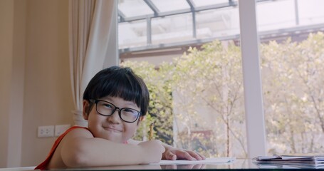 cute Asian preschool child boy with eyeglasses writing in exercise book doing homework studying at...
