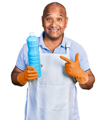 Hispanic middle age man wearing cleaner apron holding cleaning products smiling happy pointing with hand and finger
