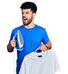 Young arab man with beard holding electric steam iron and white t shirt angry and mad screaming...