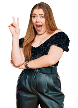 Young beautiful redhead woman wearing elegant clothes smiling with happy face winking at the camera doing victory sign. number two.