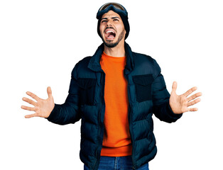 Young hispanic man with beard wearing snow wear and sky glasses crazy and mad shouting and yelling with aggressive expression and arms raised. frustration concept.
