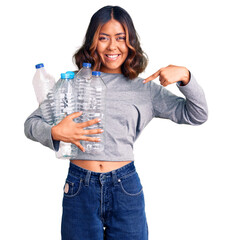 Young beautiful mixed race woman holding recycling plastic bottles pointing finger to one self...