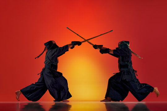 Dynamic image of two men, professional kendo athletes training with bamboo shinai sword against gradient red studio background in neon light. Concept of martial arts, sport, Japanese culture, motion