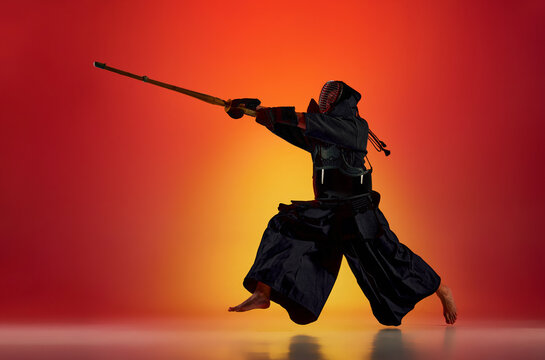 Man, professional kendo athlete in black uniform with sword, shinai training against gradient red studio background in neon light. Concept of martial arts, sport, Japanese culture, action and motion