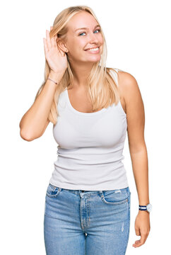 Young blonde girl wearing casual style with sleeveless shirt smiling with hand over ear listening an hearing to rumor or gossip. deafness concept.