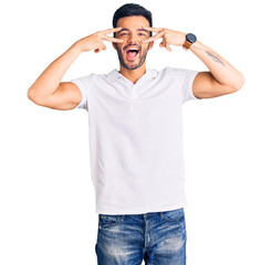 Fototapeta premium Young handsome hispanic man wearing casual clothes doing peace symbol with fingers over face, smiling cheerful showing victory