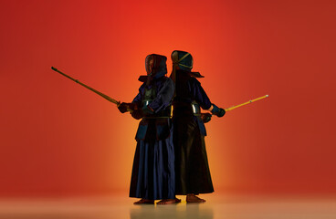 Two men, professional kendo athletes in black uniform posing with shinai sword against gradient red studio background in neon light. Concept of martial arts, sport, Japanese culture, action and motion