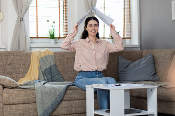 Smiling pretty young Caucasian woman sitting on sofa and holds documents over her head in form of roof. Concept of loan and mortgage