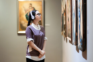 Side view of young Caucasian pretty woman wearing headphones and contemplates ancient artefacts. Student visiting arts exhibition. Concept of culture education