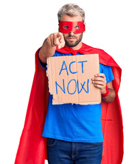 Young blond man wearing super hero costume holding act now cardboard banner pointing with finger to...
