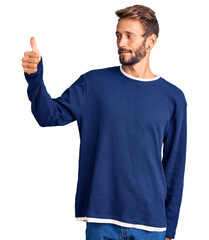 Handsome blond man with beard wearing casual sweater looking proud, smiling doing thumbs up gesture to the side