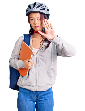 Young beautiful chinese girl wearing student backpack and bike helmet holding book with open hand doing stop sign with serious and confident expression, defense gesture